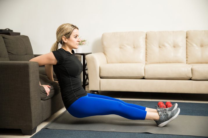 Profile view of a pretty young woman doing tricep dips leaning on a couch in her living room